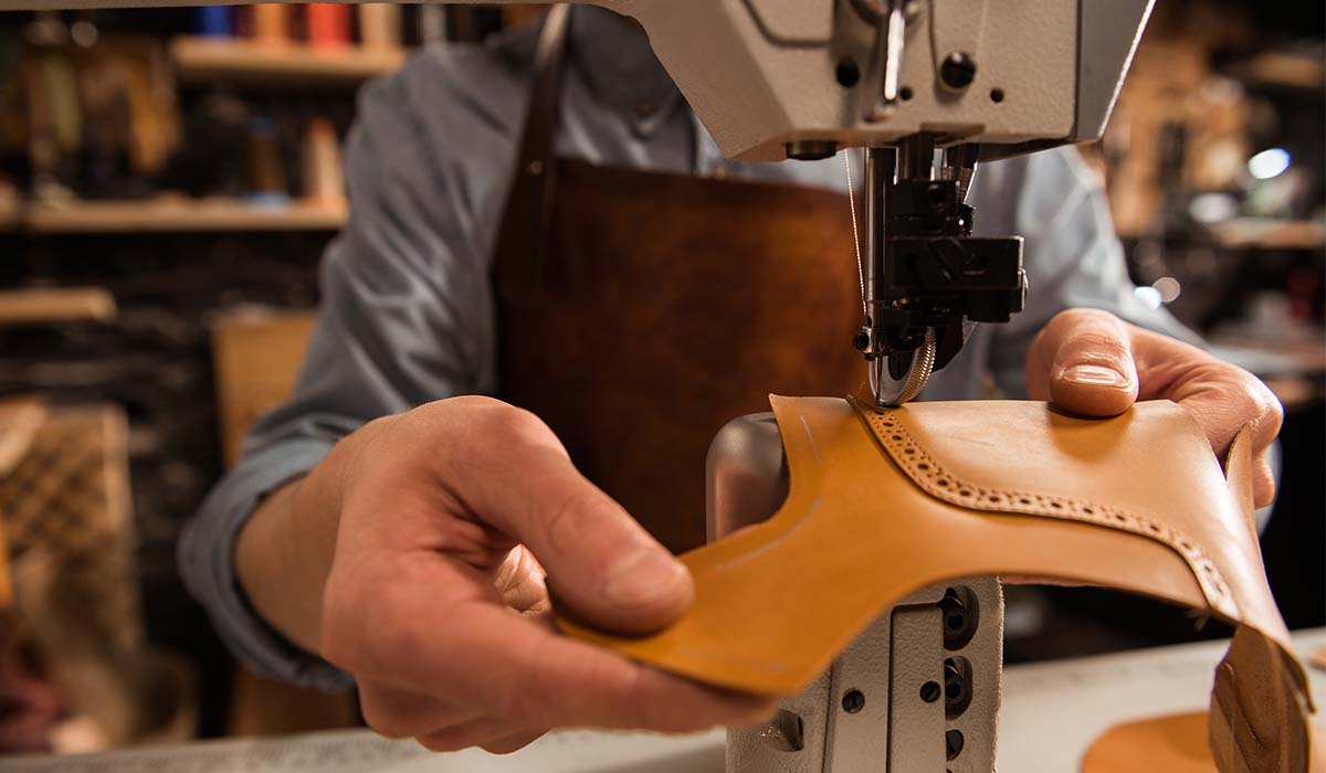 Pengrajin sepatu sedang menjahit potongan kulit dengan mesin jahit khusus dalam proses pembuatan sepatu